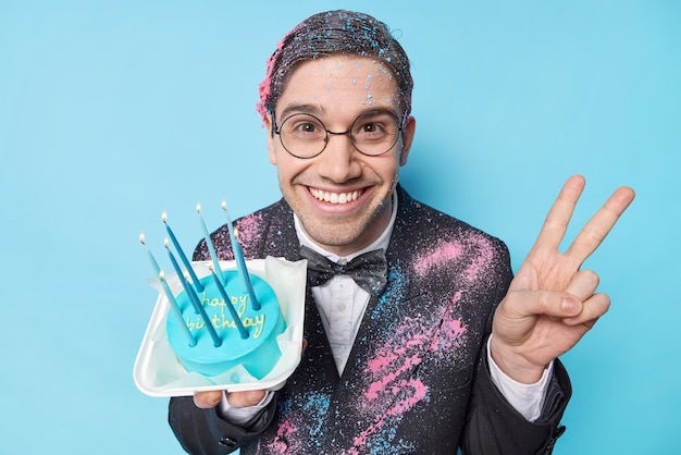 Un homme heureux heureux de célébrer son anniversaire tient un gâteau sucré avec des bougies allumées fait un geste de paix porte des lunettes rondes une veste formelle noire et un nœud papillon sourit largement isolé sur fond bleu