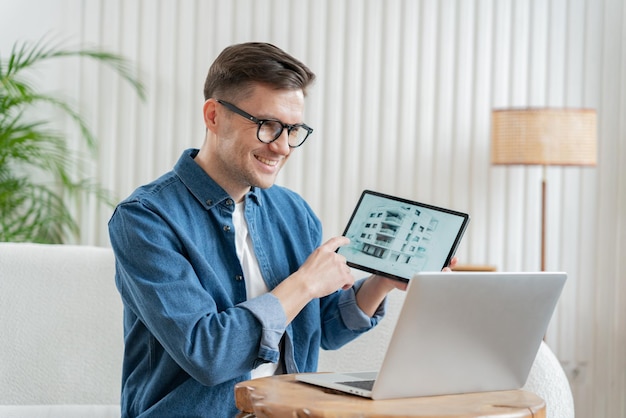 Un homme heureux en lunettes montre un écran de tablette avec un plan de bâtiment assis à côté d'un ordinateur portable dans une pièce confortable