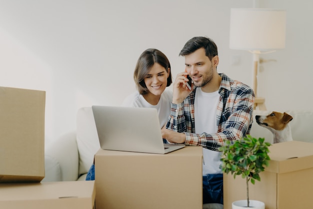 Homme heureux navigue sur un ordinateur portable dans un nouvel appartement, appelle via un smartphone, emménage dans un nouvel appartement avec sa femme