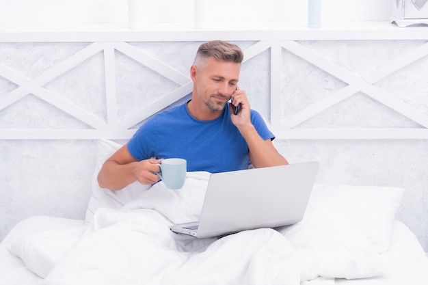 Un homme heureux parle sur son téléphone portable en buvant du café et en utilisant un ordinateur portable au lit dans la communication du matin