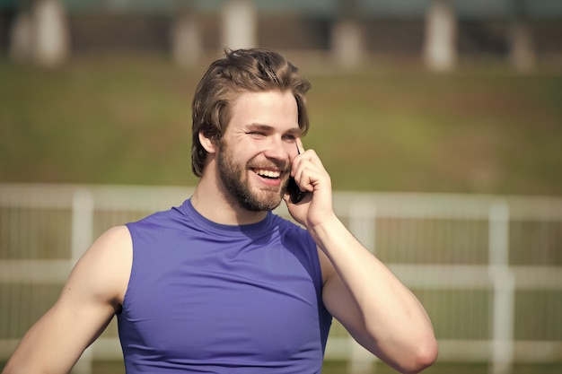 Un homme heureux parle sur un téléphone portable sur une communication extérieure ensoleillée Un homme barbu sourit avec la vie moderne d'un smartphone Sportif en mode t-shirt de sport bleu Mode sportive et mode de vie sain