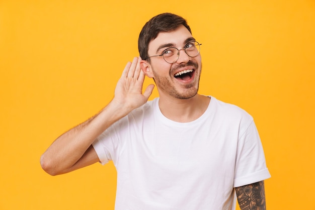 homme heureux de race blanche à lunettes posant et écoutant isolé sur un mur jaune