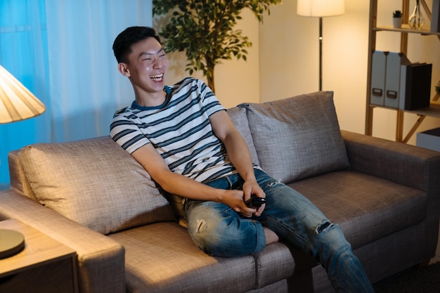 Un homme heureux regarde la télévision le soir dans le salon de la maison. jeune homme coréen asiatique assis sur un canapé et riant joyeux tout en regardant un film drôle à la télévision tard dans la nuit. un gars joyeux profite de son temps libre