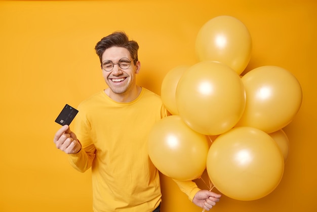 Un homme heureux rit joyeusement dépense beaucoup d'argent pour la célébration détient une carte de crédit et un tas de ballons gonflés anticipe l'anniversaire habillé avec désinvolture isolé sur fond jaune heureux d'obtenir un salaire