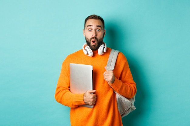 Homme heureux avec sac à dos et écouteurs, tenant un ordinateur portable et souriant, l'air surpris, debout sur fond turquoise.