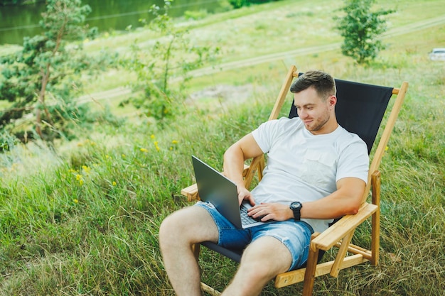 Un homme heureux se détend dans la nature et travaille à distance sur un ordinateur portable Travail à distance pendant les vacances Travail sur un ordinateur portable tout en étant assis dans une chaise dans la nature