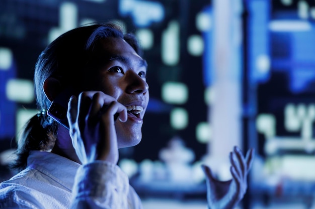 Homme heureux se promenant dans le paysage urbain pendant la nuit, appréciant la discussion au téléphone avec sa petite amie, en gros plan. Citoyen asiatique passant des appels téléphoniques pendant ses déplacements, arrière-plan flou