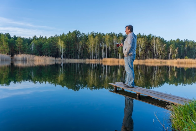 Un homme heureux se tient sur une jetée en bois avec une tasse de thé en verre près de la forêt printanière sur un lac calme en Ukraine. Concept de nature et de voyage. Scène magnifique et colorée