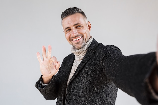 Homme heureux séduisant portant un manteau debout isolé sur un mur gris, prenant un selfie, montrant ok