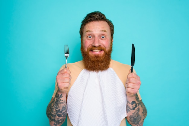 Un homme heureux avec des tatouages est prêt à manger avec des couverts à la main