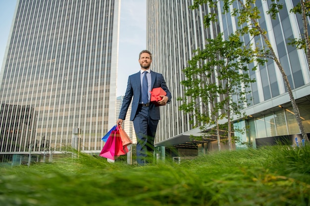 Un homme heureux en tenue de soirée tient des sacs à provisions et une boîte présente à l'extérieur de l'occasion du bureau