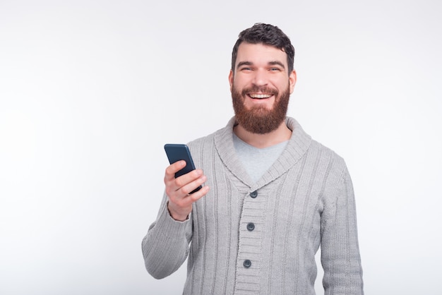 Un homme heureux tient son téléphone tout en souriant à la caméra sur fond blanc.