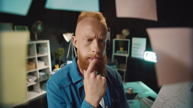 Homme hipster aux cheveux roux regardant le tableau de concept dans un bureau sombre Professionnel masculin pensif à la recherche de la bonne solution à l'intérieur Homme d'affaires concentré recherchant le bon autocollant sur le lieu de travail