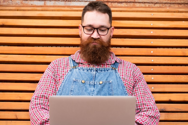 Homme hipster barbu brutal sérieux en salopette de jeans travaillant sur ordinateur portable assis sur un banc à l'extérieur. Travail à distance, travail à distance.