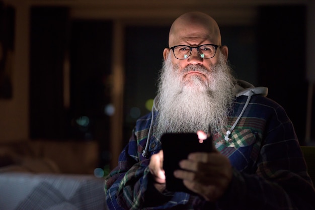 Homme hipster barbu chauve mature à l'aide de téléphone à la maison tard dans la nuit dans l'obscurité