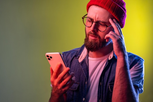 Homme hipster barbu réfléchi tenant un smartphone à la main en contemplant la mise à jour du logiciel en pensant choisir des tarifs appropriés Prise de vue en studio intérieur isolée sur fond de néon coloré