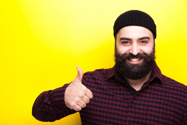Homme hipster barbu souriant avec son pouce vers le haut sur fond jaune en photo de studio