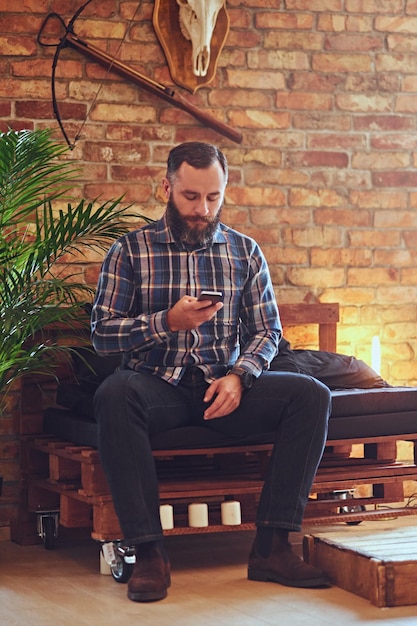 Un homme hipster barbu vêtu d'une chemise polaire à l'aide d'un smartphone dans une pièce avec un intérieur loft.