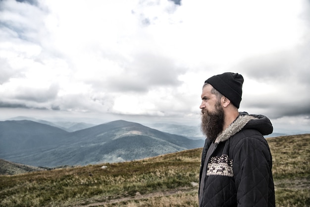 Homme hipster ou gars avec barbe et moustache sur un visage sérieux en chapeau et veste en plein air au sommet de la montagne contre ciel nuageux sur fond naturel, homme barbu