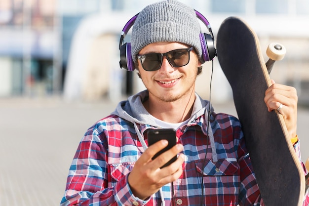 Homme hipster avec des lunettes vêtu d'une chemise à carreaux et d'une casquette tenant un smartphone et une planche à roulettes écoutant de la musique
