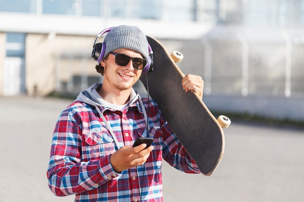 Homme hipster avec des lunettes vêtu d'une chemise à carreaux et d'une casquette tenant un smartphone et un skateboard écoutant le concept de musique