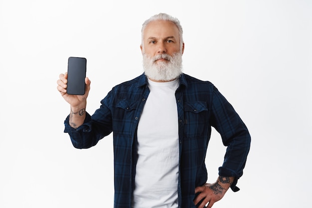 Un homme hipster mature montre l'écran d'un smartphone et a l'air confiant à l'avant, une application mobile publicitaire pour les personnes âgées, se dresse sur un mur blanc