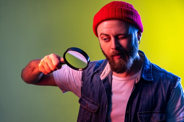 Homme hipster regardant à travers une loupe, espionnant, découvrant quelque chose, explorant et inspectant, portant un bonnet et un gilet en jean. Prise de vue en studio intérieure isolée sur fond néon coloré.