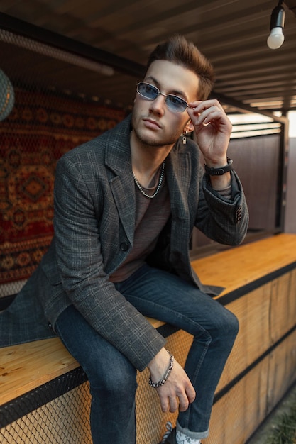 Homme hipster vogue à la mode en manteau décontracté élégant et jeans assis et porte des lunettes de soleil dans la ville