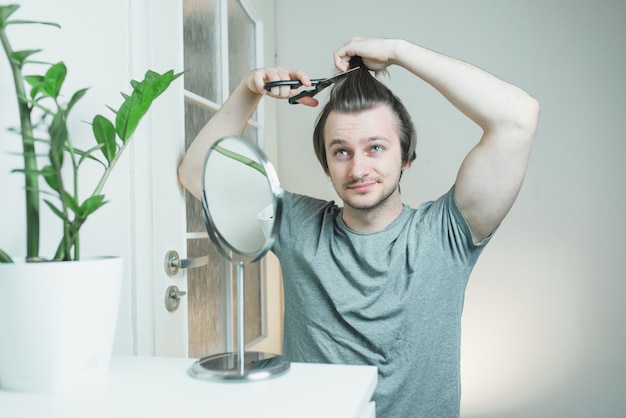Un homme hirsute en T-shirt gris avec des ciseaux dans les mains essaie de se couper les cheveux tout en se regardant dans le miroir.