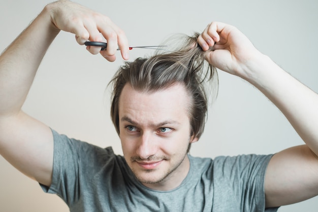 Un homme hirsute en T-shirt gris avec des ciseaux dans les mains essaie de se couper les cheveux.