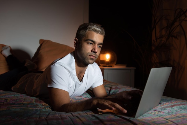 Homme hispanique allongé sur son lit et travaillant sur un ordinateur portable tard dans la nuit.