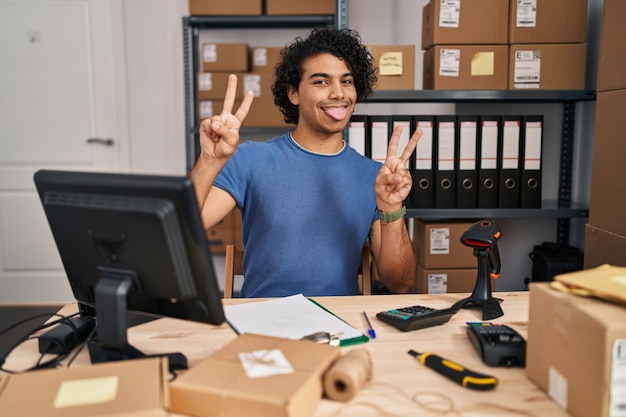 Homme hispanique aux cheveux bouclés travaillant dans une petite entreprise de commerce électronique souriant avec la langue montrant les doigts des deux mains faisant le signe de la victoire. numéro deux.