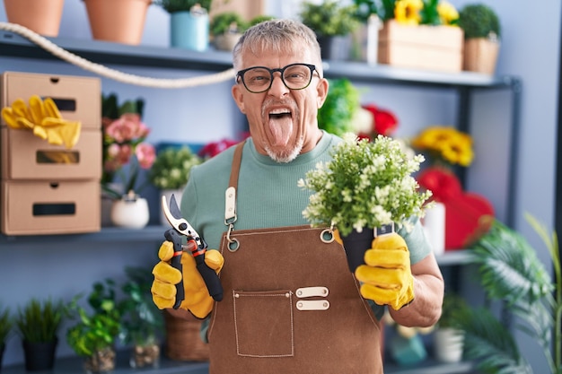 Homme hispanique aux cheveux gris travaillant dans un magasin de fleuriste qui sort la langue heureux avec une expression drôle