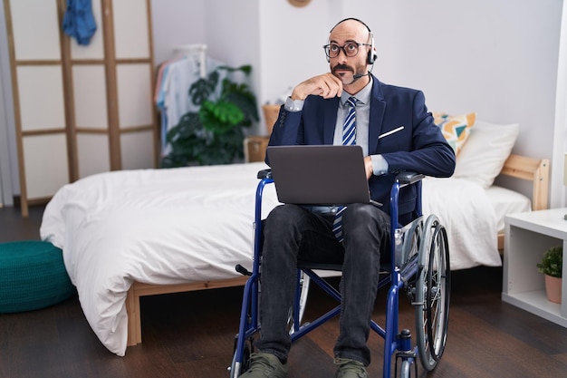 Homme hispanique avec barbe assis sur un fauteuil roulant faisant un appel vidéo d'affaires visage sérieux pensant à la question avec la main sur le menton, pensif à l'idée confuse