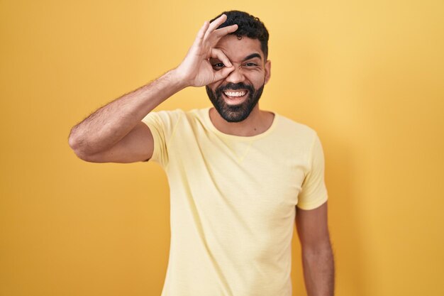 Homme hispanique avec barbe debout sur fond jaune faisant un geste correct avec la main souriante, les yeux regardant à travers les doigts avec un visage heureux.