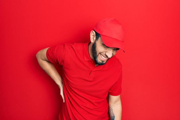 Homme hispanique avec barbe portant un uniforme de livraison et une casquette souffrant de maux de dos touchant le dos avec des douleurs musculaires à la main