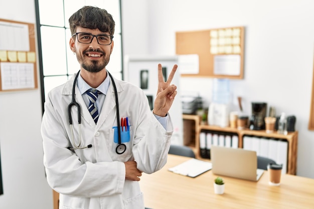 Homme hispanique avec barbe portant un uniforme de médecin et un stéthoscope au bureau souriant avec un visage heureux faisant un clin d'œil à la caméra faisant le signe de la victoire. numéro deux.