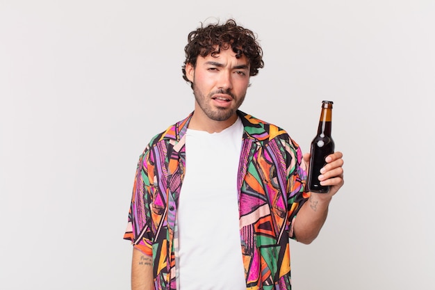 Homme hispanique avec de la bière se sentant perplexe et confus, avec une expression stupide et stupéfaite en regardant quelque chose d'inattendu