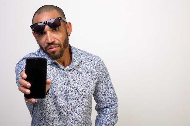 Un homme hispanique cool avec des lunettes de soleil sur le front tient un téléphone portable avec un écran vide sur fond blanc