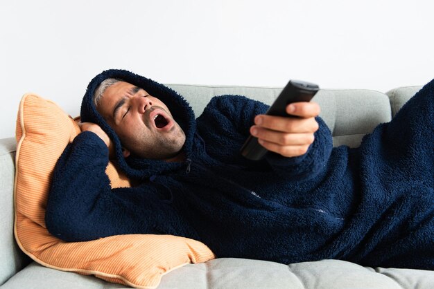 Homme hispanique ennuyé portant un pyjama d'hiver surfant sur les chaînes et bâillant tout en étant allongé sur un canapé.
