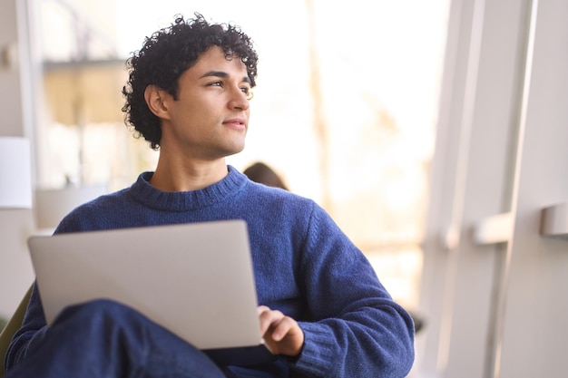 Homme hispanique pensif regardant pensivement de côté tout en travaillant en ligne sur un ordinateur portable depuis un lieu de coworking