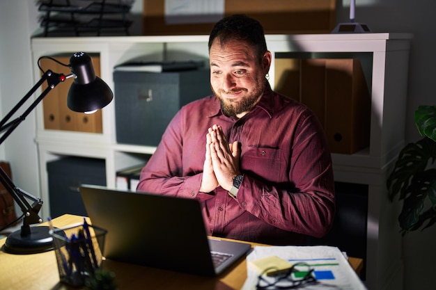 Homme hispanique de taille plus avec barbe travaillant au bureau la nuit priant les mains jointes pour demander pardon souriant confiant