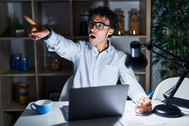 Homme hispanique travaillant au bureau la nuit pointant du doigt surpris devant la bouche ouverte expression étonnée quelque chose sur le devant