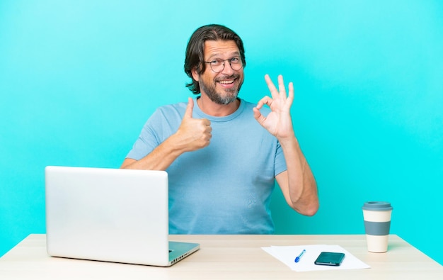 Homme hollandais senior dans une table avec un ordinateur portable isolé sur fond bleu montrant le signe ok et le geste du pouce vers le haut