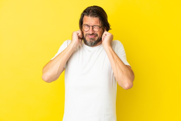 Homme hollandais senior isolé sur fond jaune frustré et couvrant les oreilles