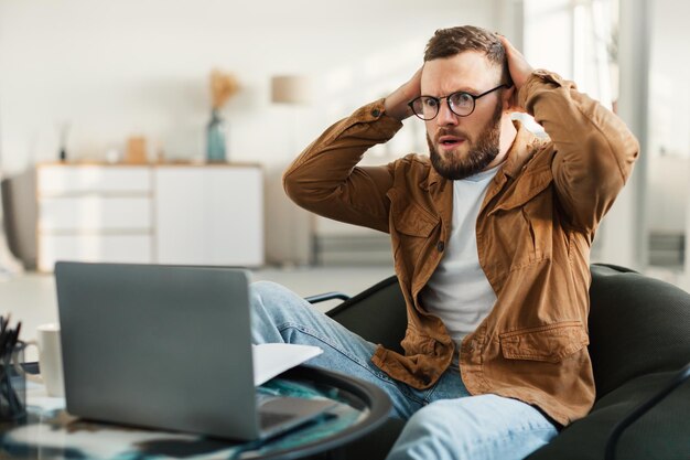 Homme indépendant choqué regardant un ordinateur portable touchant la tête à la maison