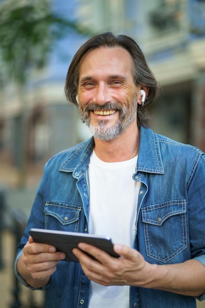 Homme indépendant confiant avec une tablette numérique aux cheveux gris dans les mains ayant un appel tout en se tenant dans les rues urbaines Homme mûr parlant ou écoutant de la musique utiliser des écouteurs sans fil voyager dans la vieille ville