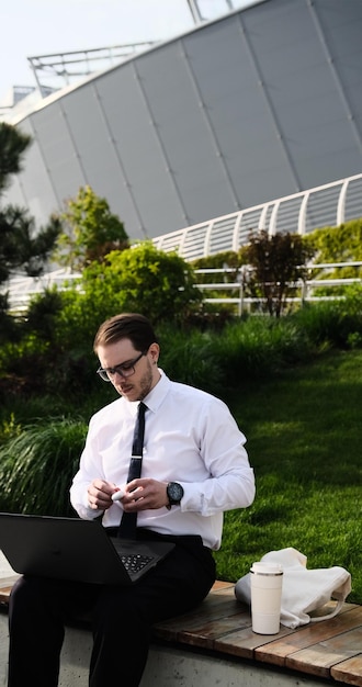 homme indépendant travaillant à distance en ligne avec des écouteurs et un ordinateur portable conférence d'appel vidéo ou réunion à distance homme dans un parc de la ville sur un banc un fond urbain moderne à l'extérieur en plein air