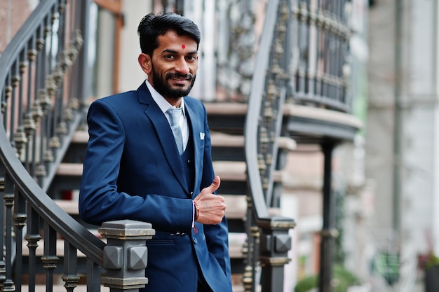 Homme indien barbe élégante avec bindi sur le front porter sur costume bleu posé en plein air contre des escaliers en fer et montrer le pouce vers le haut