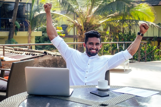 Homme indien beau et réussi dans un pigiste élégant et bien habillé travaillant avec un ordinateur portable sur la plageétudiant indépendant et à distance dans un café d'été sur la rive de l'océan indien